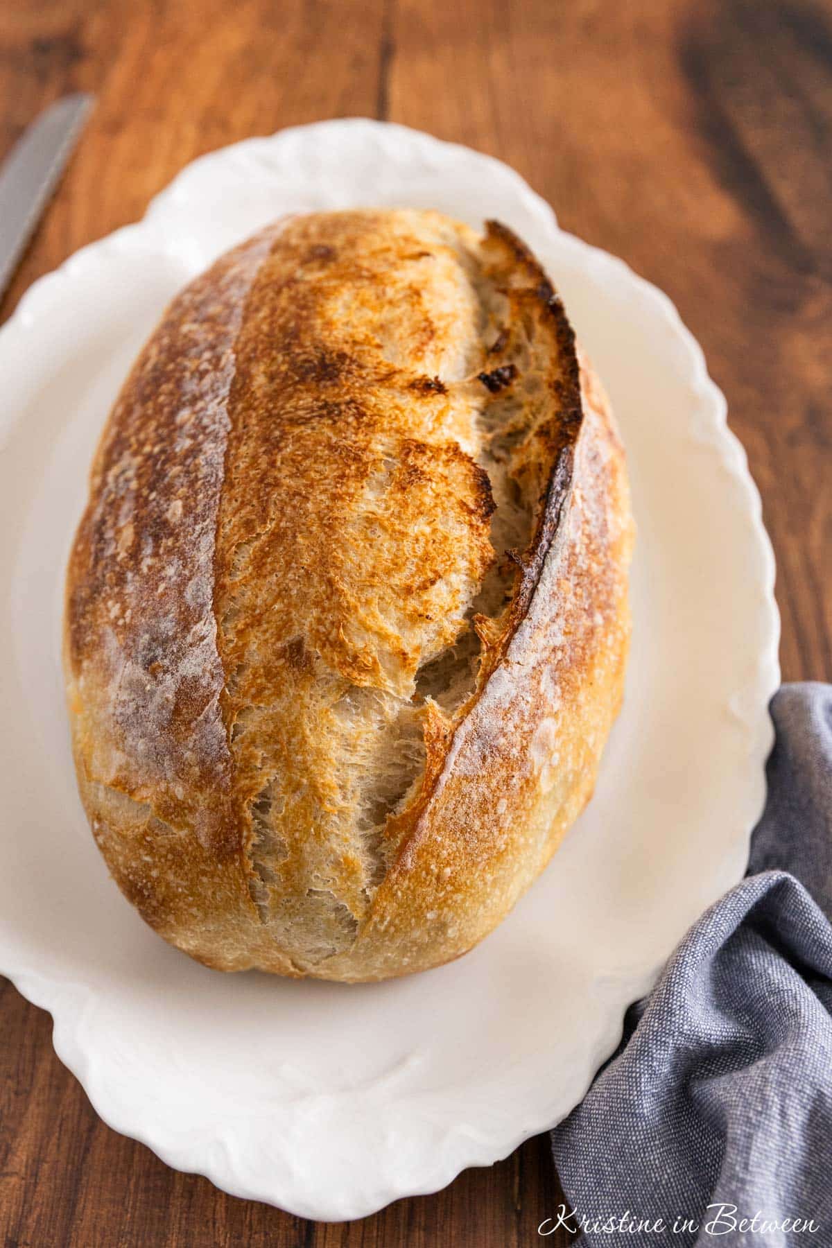 A whole loaf of sourdough bread on a white platter with a blue napkin sitting next to it.