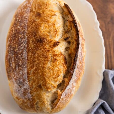 A whole loaf of sourdough bread on a white platter with a blue napkin sitting next to it.