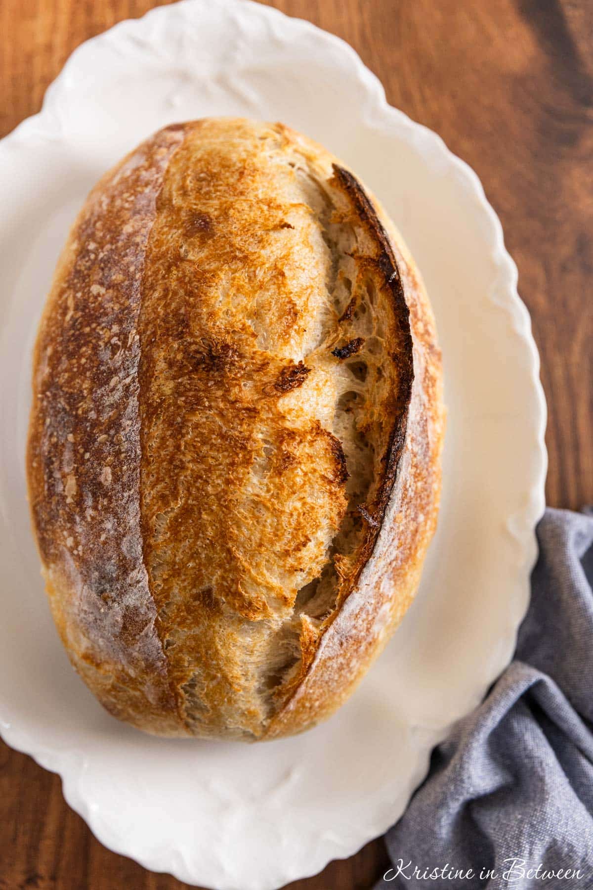 A whole loaf of sourdough bread on a white platter with a blue napkin sitting next to it.